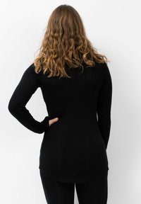 Persoon met golvend lichtbruin haar, gekleed in een zwart shirt met lange mouwen en zwarte broek, staande met één hand op de heup, met de rug naar de kijker gekeerd.