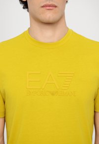 Geltona medvilninė marškinė su tekstūruotu EA7 Emporio Armani logotipu priekyje, apvali iškirptė ir trumpomis rankovėmis.