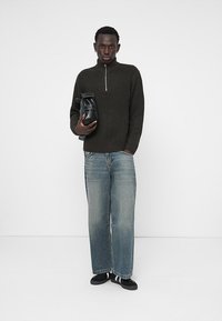 Dark green knit sweater with a half-zip design, paired with light blue loose-fitting jeans and black slip-on shoes. Holding a black bag.
