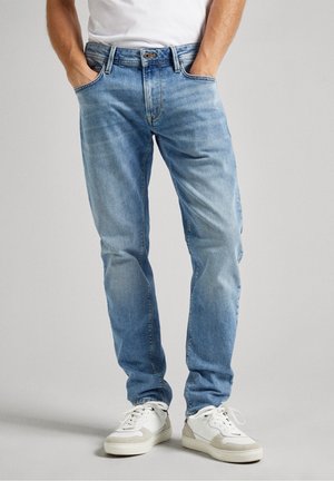 Homme portant un jean bleu clair, mains dans les poches et baskets blanches sur un fond gris uni.