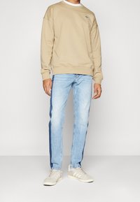 Ljusbeige sweatshirt med rund halsringning och subtil logotyp, kombinerad med ljusblå jeans med mörka sidostreck och ljusa sneakers.