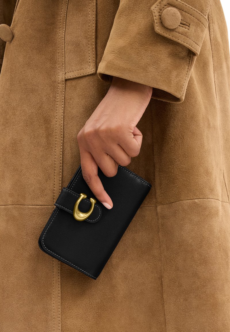 Main tenant un portefeuille en cuir noir avec une fermeture dorée contre la manche et le corps d'un manteau en daim marron.