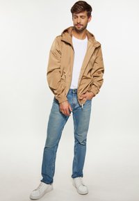 Beige lättviktsjacka med huva och elastiska manschetter, parat med blå jeans och vita sneakers. Enkel design med en avslappnad passform.