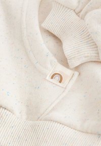 Kremasta pletena tkanina z večbarvnimi pegicami, izvlečeni manšetni rob in majhna etiketa z minimalističnim dizajnom mavrice.