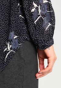 Blusa navy con pois bianchi e motivo floreale in diverse tonalità di blu. Presenta maniche lunghe e polsini con bottoni. Tessuto ruvido.