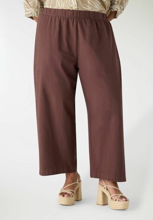 Donna che indossa pantaloni marroni a gamba larga e sandali con zeppa beige con cinturini alla caviglia su sfondo bianco.