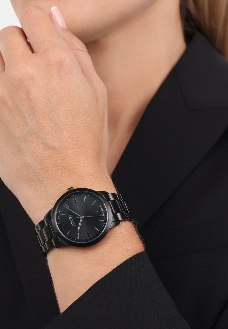 Zwarte metalen horloge met een ronde wijzerplaat, zilveren uurmarkeringen en een schakelarmband. Draagt op een pols tegen een zwarte blazer achtergrond.