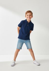 Granatowa koszulka polo z trzema guzikami, w zestawieniu z jasnymi szortami dżinsowymi. Dziecko stoi z rozstawionymi nogami, nosząc białe trampki.