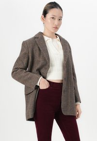 Ung kvinne iført en brunrutete blazer, en kremfarget, kabelstrikket, kort genser og burgunderhøye bukser med den ene hånden i lommen.