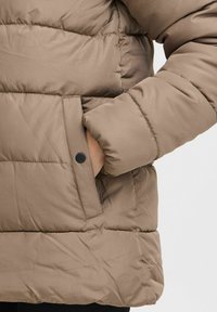 Veste en duvet beige avec une texture matelassée, dotée d'une poche latérale avec un bouton-pression noir et d'une silhouette décontractée.