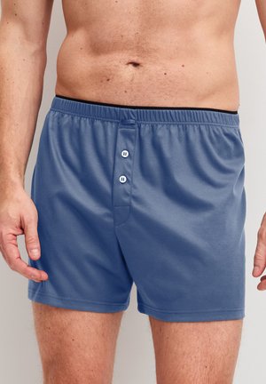 Uomo che indossa boxer di cotone blu con due bottoni bianchi sul davanti, mostrando il torso e la parte superiore delle cosce su uno sfondo uniforme.