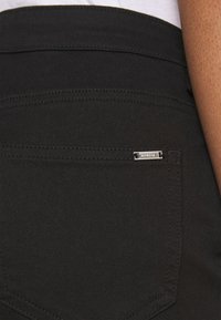 Jeans de mezclilla negra con un primer plano del bolsillo trasero, que presenta una etiqueta de metal para la marca y detalles de costura visibles.
