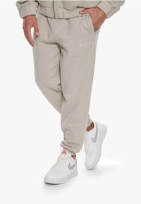 Beige jogger pants with elastic waistband, side pockets, and "Dropside" embroidered detail on the thigh. Paired with white sneakers.