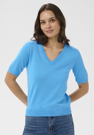 Femme aux cheveux mi-longs et foncés portant un pull bleu vif à manches courtes avec un col en V et un jean bleu, debout devant un fond clair uni.