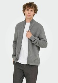 Grijze ritsvest met textuurpatroon, hoge kraag en geribde manchetten, gedragen over een wit T-shirt, in combinatie met donkere, gestructureerde broeken.
