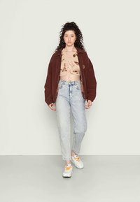 Casaco oversized castanho, top bege com padrões castanhos, jeans azul claro e ténis brancos com atacadores amarelos. Design simples e ajuste descontraído.