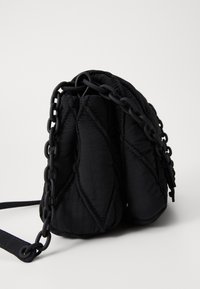 Schwarze, gesteppte Stoff-Umhängetasche mit dickem schwarzen Kunststoffkettenriemen, vor einem schlichten hellen Hintergrund aus der Seitenansicht präsentiert.