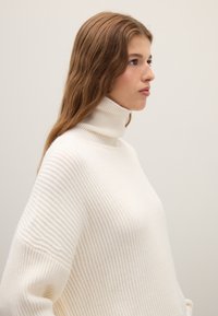 Weißer gerippter Rollkragenpullover mit entspanntem Sitz und weicher Textur, ausgestattet mit einem hohen Kragen und breiten Ärmeln für Komfort.
