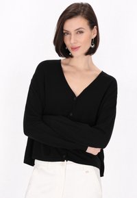 Černý cardigan s výstřihem do V, který má volný střih a tři knoflíky vpředu. Měkká textura a krátké široké rukávy.