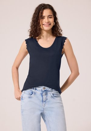 Femme souriante aux cheveux bouclés portant un haut noir sans manches avec une bordure en dentelle et un jean en denim bleu clair sur un fond uni.