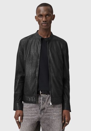 Homme portant une veste en cuir noire sur une chemise noire et un jean gris délavé, debout devant un fond gris uni.