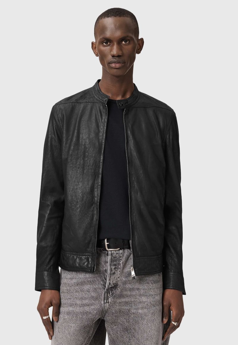 Άνδρας που φοράει μαύρο δερμάτινο μπουφάν πάνω από ένα μαύρο πουκάμισο και ξεβαμμένα γκρι τζιν, στέκεται μπροστά σε απλό γκρι φόντο.