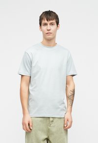 Ανοιχτό γκρίζο κοντομάνικο μπλουζάκι με στρογγυλή λαιμόκοψη, φτιαγμένο από απαλό ύφασμα. Συνοδεύεται από πράσινους τζιν και ένα ορατό τατουάζ στον βραχίονα.