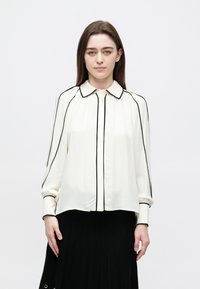Woman with straight brown hair wears a white blouse with black piping and a black pleated skirt, standing against a plain white background.