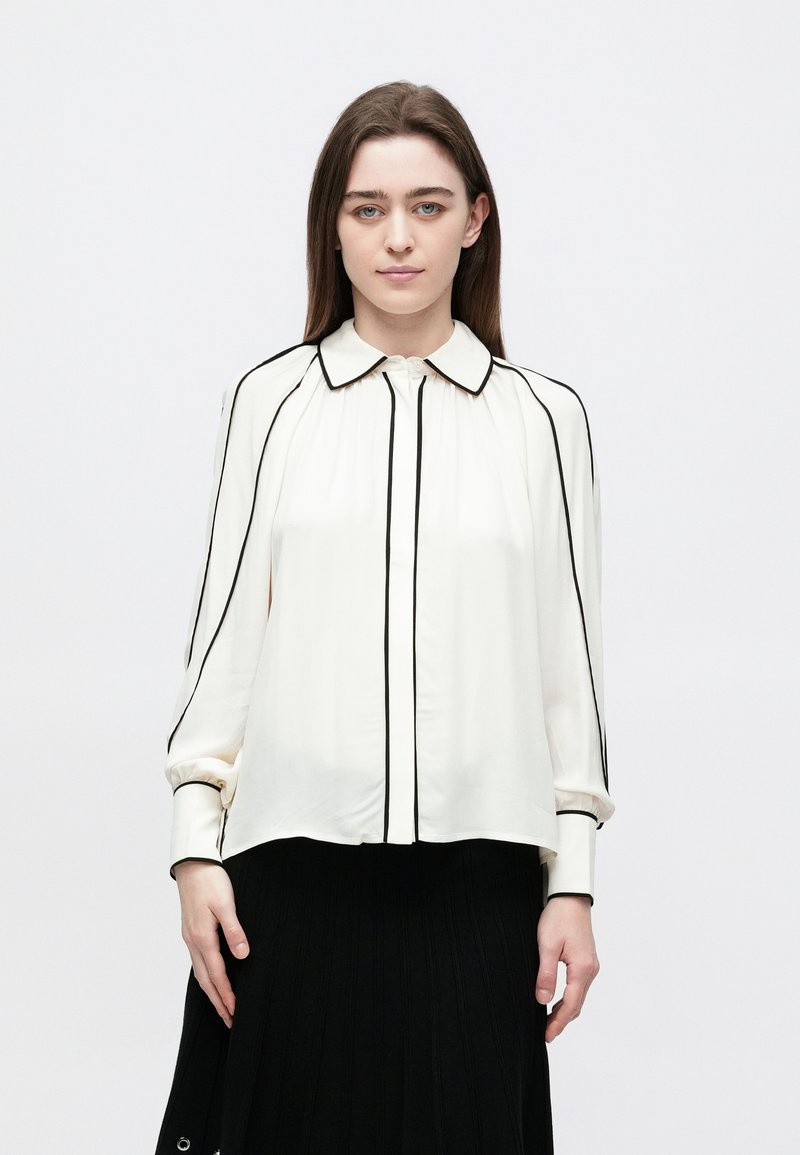 Woman with straight brown hair wears a white blouse with black piping and a black pleated skirt, standing against a plain white background.