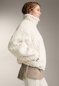 Přehnaně velká krémová fleecová bunda s vysokým límcem, texturovaným vzorem a postranními rozparky. Doplněna béžovými pletenými akcenty na spodním lemu.