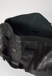 Sac de sport noir en matériau texturé avec des motifs géométriques. Dispose de poignées doubles et d'une fermeture éclair révélant un intérieur vert.