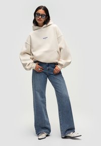 Helle beige Oversize-Hoodie mit einer Vordertasche, versehen mit blauem Logotext. Kombiniert mit hoch taillierten, geraden blauen Jeans und weißen Schuhen.