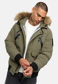 Olivgrüner Pufferjacke mit einer mit Fell besetzten Kapuze, zwei Fronttaschen, gerippten Bündchen und einem silbernen Innenfutter. Verstellbarer Kordelzug in der Taille.