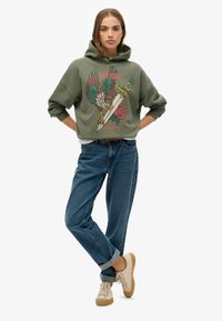 Hoodie vert olive avec imprimé graphique présentant une épée, un oiseau et des roses. Assorti à un jean bleu à ourlet roulé et des baskets beige clair.