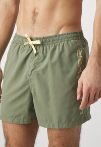 Groene zwembadschoenen van lichtgewicht materiaal, met een elastische tailleband en een gele koord, en een kleurrijk logo-ontwerp aan de zijkant.