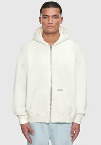 Hvid zip-op hættetrøje med en frontlomme, oversized pasform, justerbar hætte og ribbede manchetter. Blødt stof med en glat tekstur.