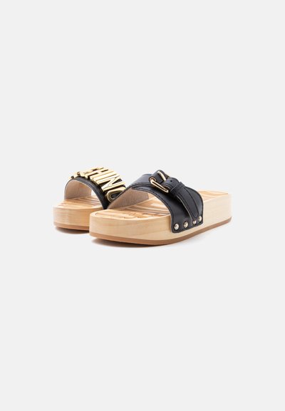 Sandales à plateforme en bois avec des brides en cuir noir ornées d'une boucle dorée et de lettres dorées, sur fond blanc.