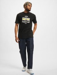Sort t-shirt med grafiktryk, parret med mørke denimbukser med en lomme. Skoene er hvide med blå accenter.