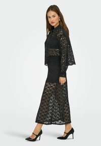 Robe en dentelle noire avec des manches longues, un col et une jupe présentant une fente sur le côté. Associée à des chaussures à talons hauts noires.