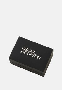 Musta suorakulmainen laatikko, jossa on sileä pinta, ja joka sisältää hopeanvärisen "OSCAR JACOBSON" -logon päällä.