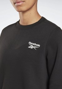 Sudadera negra de algodón, con cuello redondo y puños acanalados. Presenta un logo blanco de Reebok en el pecho izquierdo.