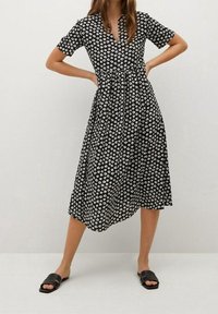 Vestido negro con patrón floral blanco, mangas cortas, escote en V, cintura ajustada y una falda A-line fluida. Combinado con sandalias negras de tipo deslizante.