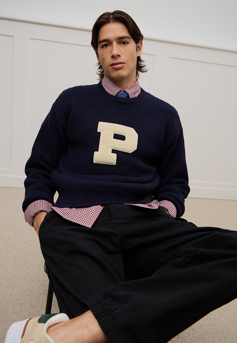 Marineblauer Strickpullover mit großem beigen "P"-Logo, getragen über einem karierten Hemd mit sichtbarem Kragen, kombiniert mit schwarzen Hosen.