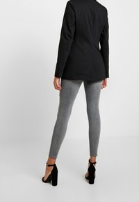 Blazer nero su misura, leggings grigi elasticizzati e scarpe con tacco alte nere. Tessuto morbido, vestibilità slim e dettaglio con cinturino alla caviglia sulle scarpe.