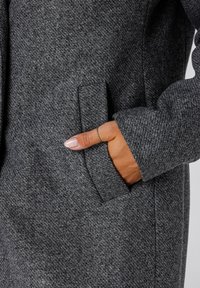 Hand met zilveren ring en lichtroze nagellak in de zak van de mouw van een donkergrijze gestructureerde wollen jas.