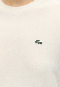 Gros plan sur une personne portant une chemise blanche texturée avec un petit logo crocodile vert sur la partie gauche de la poitrine.
