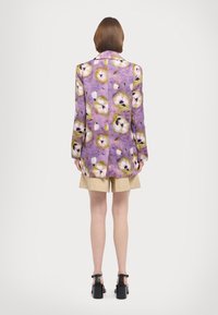 Blazer med blomstermønster i lilla og grøn, med en glat tekstur og lange ærmer. Har en spids krave og en ventileret ryg.