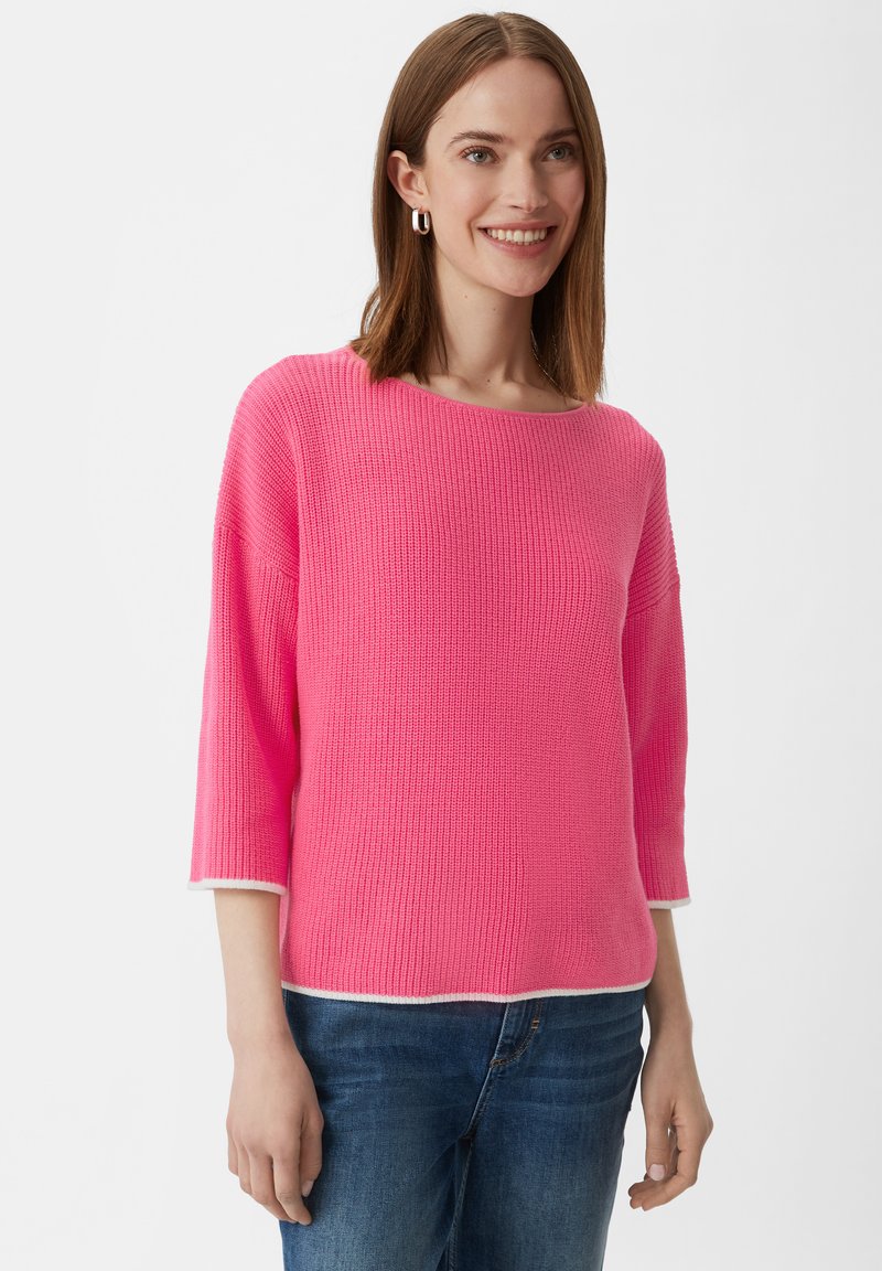 Rosa gestrickter Pullover mit Dreiviertelärmeln, weitem Ausschnitt und weißen Akzenten am Saum und an den Ärmeln, kombiniert mit blauen Jeans.