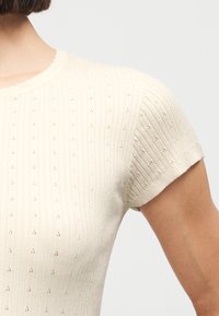 Κοντινό πλάνο ενός ατόμου που φοράει ένα πλεκτό τοπ σε εκρού χρώμα με κοντά μανίκια, διακριτικό μοτίβο με μικρές τρύπες και υφή με ριμπ.