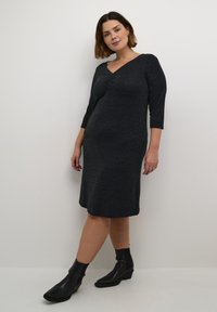 Robe noire texturée, longueur genou, avec un décolleté en V et des manches trois-quarts. Portée avec des bottines noires, se tenant contre un fond blanc.